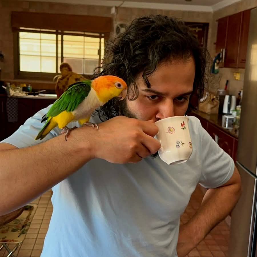 Moazzam Ullah drinking tea with a colourful parrot perched on his hand